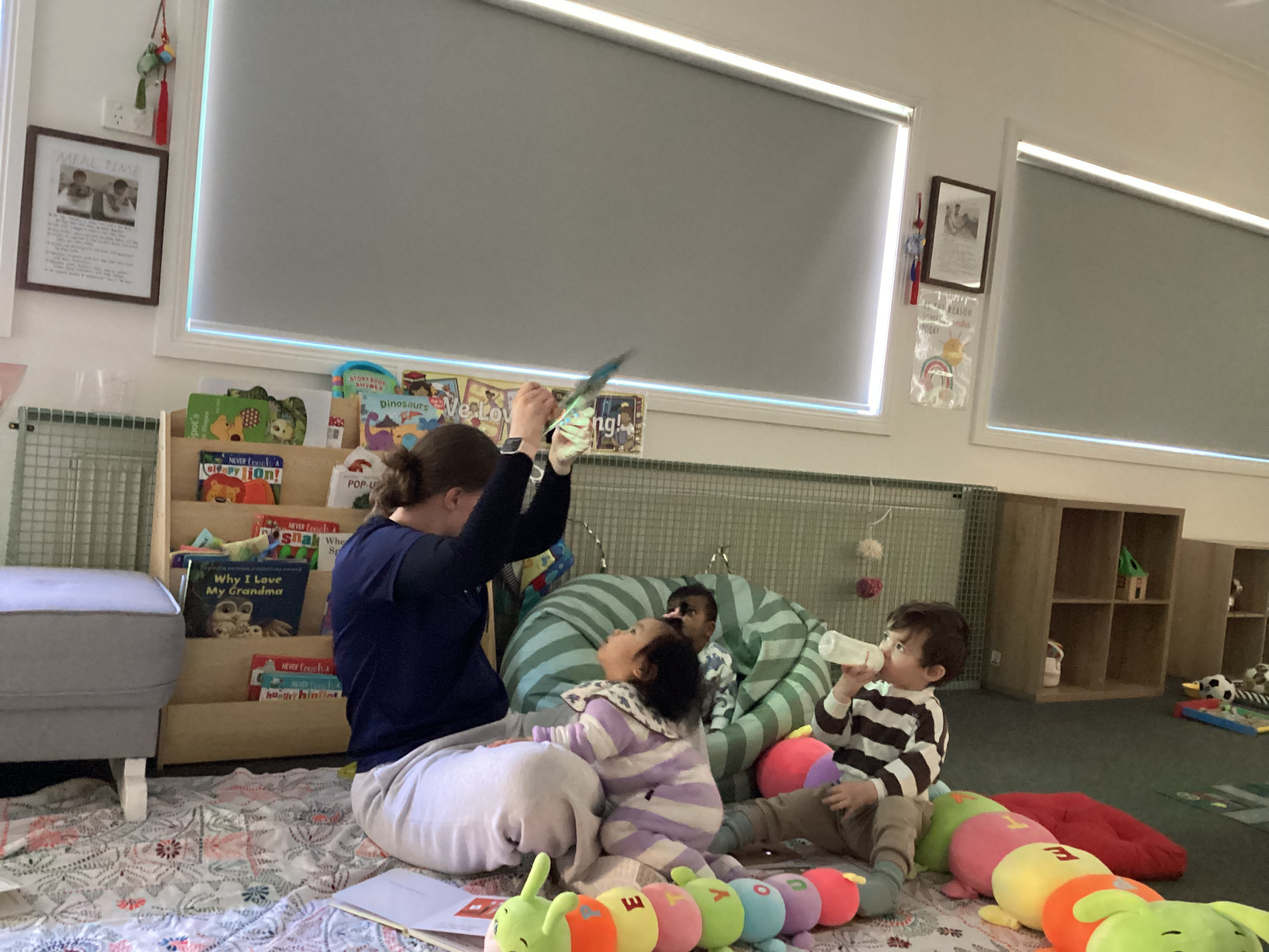 An educator sharing a story with babies in a cozy nursery corner surrounded by books and soft toys