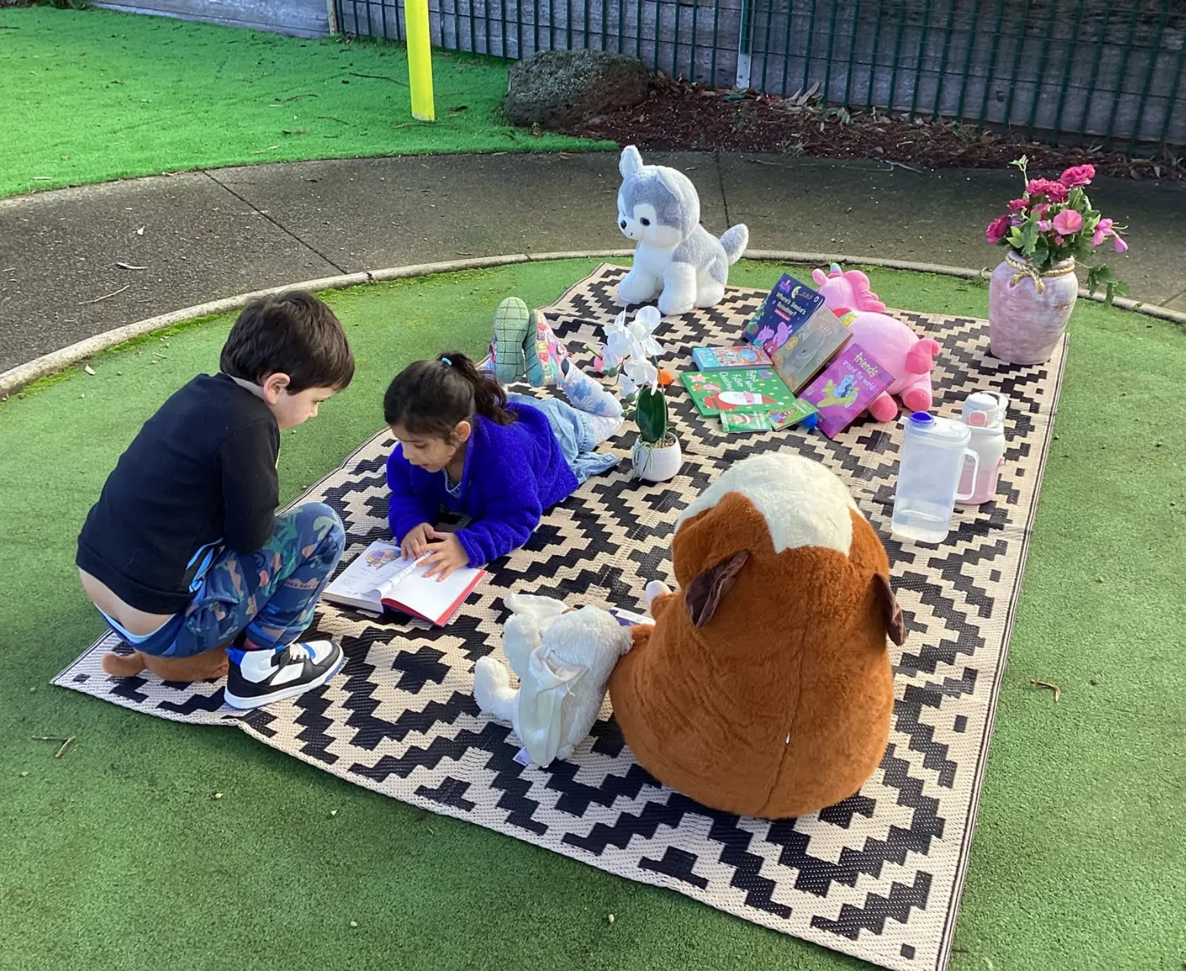 Two children reading picture books on an outdoor mat with soft toys